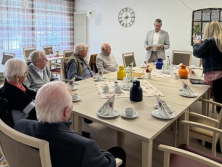 Fünf Senioren, die Gründungsmitglieder des Winterberger Stadtrats waren, sitzen am Tisch, Bürgermeister Beckmann steht und hat etwas in der Hand, über das er redet.