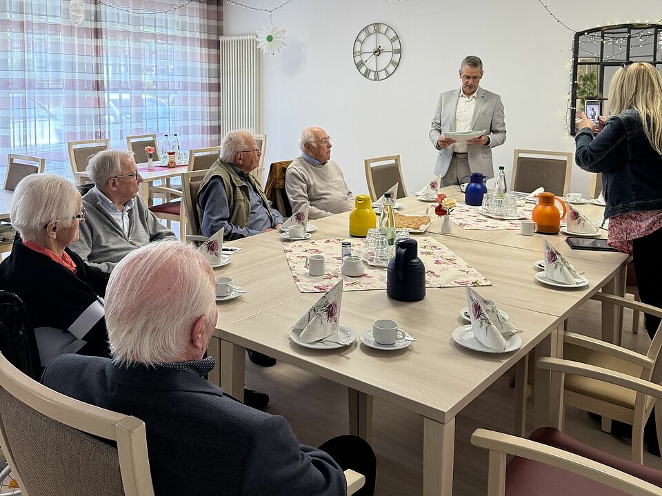Fünf Senioren, die Gründungsmitglieder des Winterberger Stadtrats waren, sitzen am Tisch, Bürgermeister Beckmann steht und hat etwas in der Hand, über das er redet.