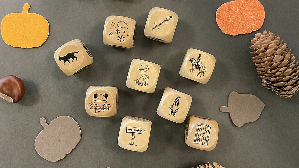 Es sind sieben Würfel mit mythischen und alltagsgebräuchlichen Wesen und Gegenständen zu sehen. Drumherum wurde herbstlich dekoriert mit Tannenzapfen, aus Pappe herausgeschnittenen Kürbissen und Nüssen und Kastanien befinden sich dort.