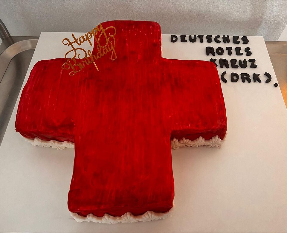 Ein Kuchen in Form eines Roten Kreuzes steht auf einem Tisch. In dem Kuchen steckt ein Nadel mit dem Schriftzug Happy Birthday. Dabenen steht mit Dekorbuchstaben "Deutsches Rotes Kreuz (DRK)":