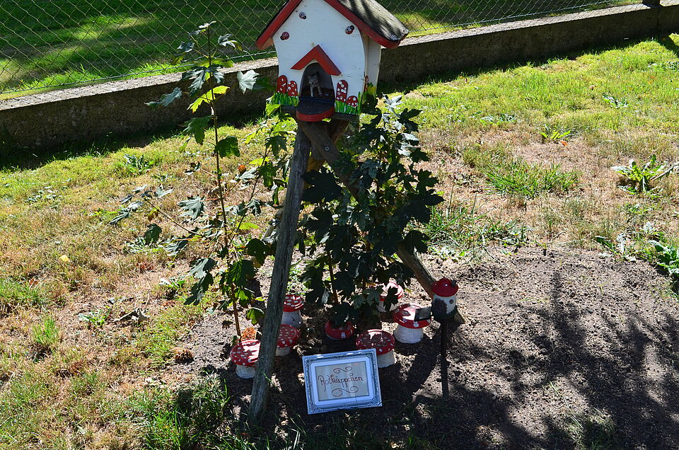 Kleine Garteninstallation mit einem weißen Häuschen auf Holzstelzen, dekoriert mit roten Fliegenpilzfiguren und grünen Pflanzen. Davor steht ein Bilderrahmen mit der Aufschrift ‚Rotkäppchen‘