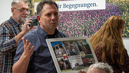 Ein Mann aus der Ukraine zeigt den Bewohnern auf einem Plakat, wie eine Senioreneinrichtung in der Ukraine aussieht. Darauf steht "Pflegeheim Yosypivka - Lebensqualität erhalten, in Würde sterben."