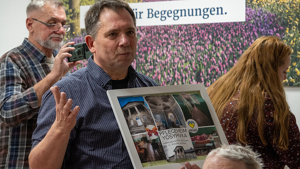 Ein Mann aus der Ukraine zeigt den Bewohnern auf einem Plakat, wie eine Senioreneinrichtung in der Ukraine aussieht. Darauf steht "Pflegeheim Yosypivka - Lebensqualität erhalten, in Würde sterben."