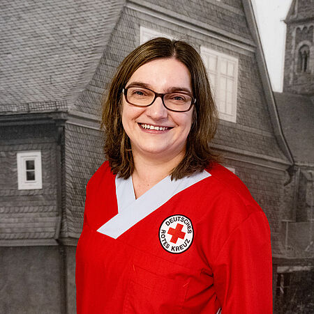 Theresa Braune Die Person auf dem Bild trägt eine rote Arbeitskleidung mit dem Logo des Deutschen Roten Kreuzes. Sie hat schulterlanges, braunes Haar und eine Brille. Sie lächelt freundlich in die Kamera. Im Hintergrund ist ein Bild eines Hauses mit Schieferfassade zu sehen, das schwarz-weiß dargestellt ist.
