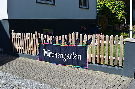 Ein Holzlattenzaun mit einem schwarzen Stoffbanner, auf dem in weißer Schrift „Märchengarten“ steht. Das Banner ist mit einer bunten Kante verziert. Dahinter ist ein Vorgarten mit Blumen und Rasen zu sehen.