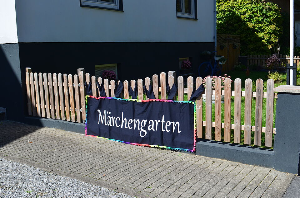 Ein Holzlattenzaun mit einem schwarzen Stoffbanner, auf dem in weißer Schrift „Märchengarten“ steht. Das Banner ist mit einer bunten Kante verziert. Dahinter ist ein Vorgarten mit Blumen und Rasen zu sehen.