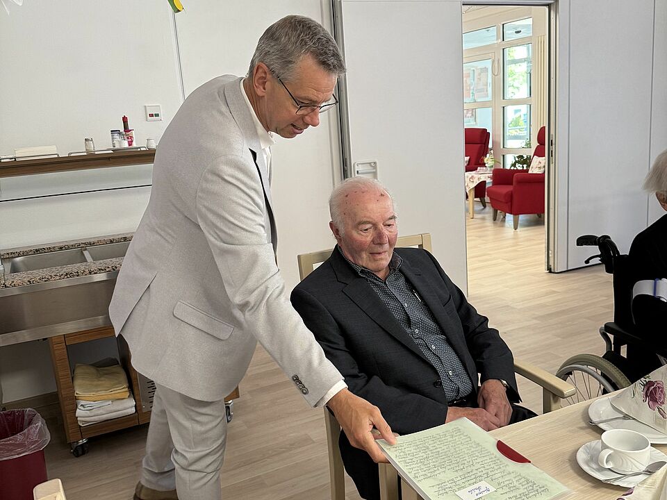 Bürgermeister Beckmann steht neben einem sitzenden Gründungsmitglied und zeigt ihm ein historisches Dokument.