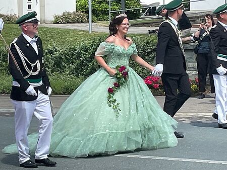 Der Schützenkönig und die Königin laufen Hand in Hand auf der Straße im Schützenzug. Die Königin hat ein hellgrünes Kleid mit einem Reifrock an.