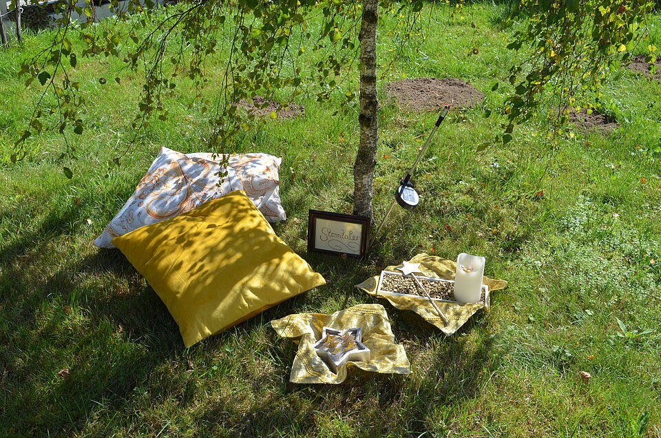 Ein gemütlicher Platz im Gras unter einem Baum. Zwei Kissen liegen auf dem Boden, daneben eine goldene Decke mit Deko-Sternen, eine Kerze und ein Zauberstab. Ein kleines Schild mit der Aufschrift „Sternsalaer“ lehnt am Baum.